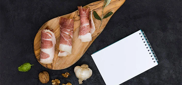 Blank white spiral notepad with rolled bacon on serving wooden board with basil; walnuts and garlic