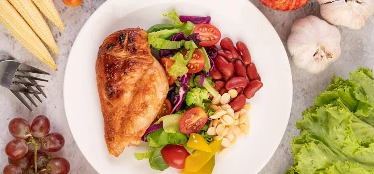 Chicken steak topped with white sesame, peas, tomatoes, broccoli and pumpkin on a white plate.