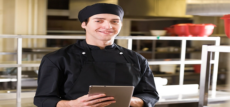 Chef wearing black apron and cap holding a tablet.