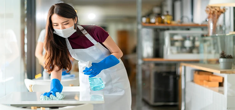 Waitress wearing face mask, apron, and rubber glove cleaning table with disinfectant. 